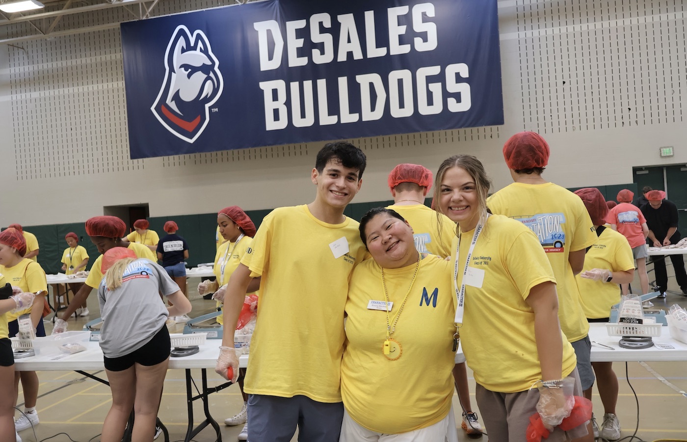 Three first-year students smiling in front of a DeSales Bulldogs banner in a room full of other first-year student volunteers.